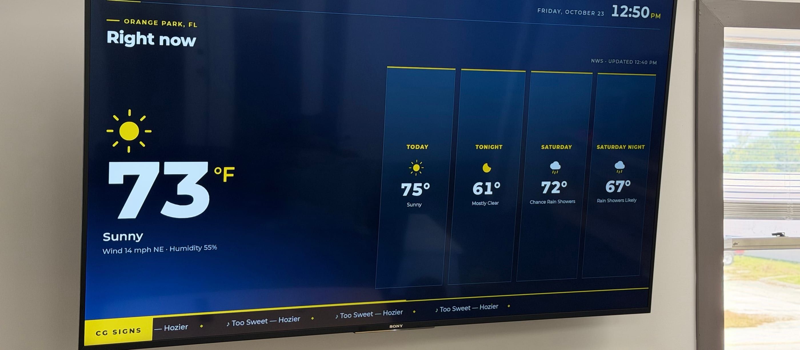 Office TV running the signage: the current weather for Orange Park, a four-part forecast, the date and time, and a ticker showing the song playing in the office.